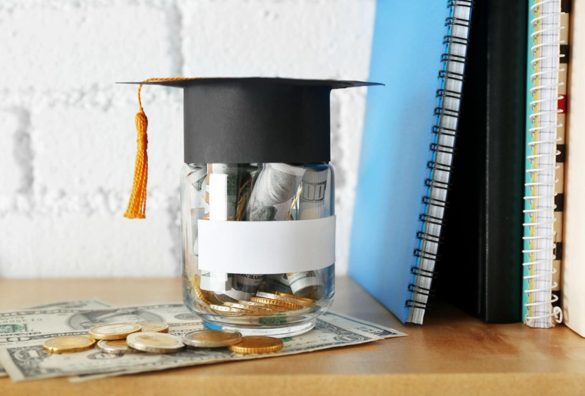 Glass jar filled with rolled dollar bills and coins wearing a graduation cap, placed beside notebooks — symbolizing saving money for vet school applications. Vetucore LLC supports future veterinarians with expert financial planning advice.